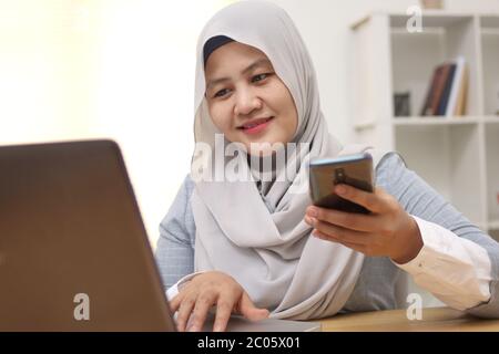 Donna musulmana asiatica lavora in ufficio, utilizzando telefono e laptop, felice espressione sorridente del viso, acquisto online check out Foto Stock
