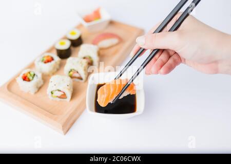 Primo piano di donna che tiene i chopsticks con il rotolo di sushi di salmone e ciotola con salsa di soia sul tavolo, porzione di pasto tradizionale giapponese in background. Foto Stock