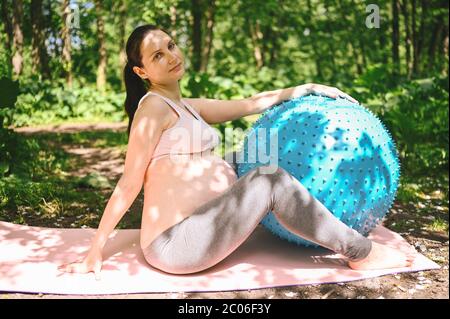 Bella giovane donna incinta che fa esercizio con fitness Pilates palla blu nel parco all'aperto. Seduta e relax su un tappetino rosa per yoga. Futuro attivo Foto Stock