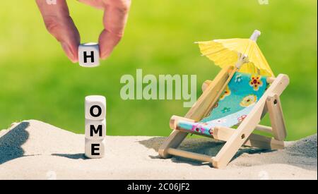 Simbolo per le vacanze durante la crisi della corona. I dadi formano la parola 'casa' accanto ad una sedia della spiaggia. Foto Stock