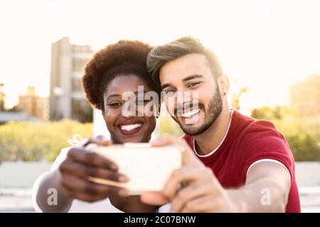 Felice coppia che prende selfie con smartphone mobile outdoor - amici multirazziali divertirsi con nuove app sociali alla moda Foto Stock