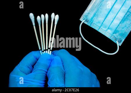 Un primo piano di mani che indossano guanti ad un medico personale che tiene i tamponi della gola accanto a una maschera medica viso su uno sfondo nero. Foto Stock