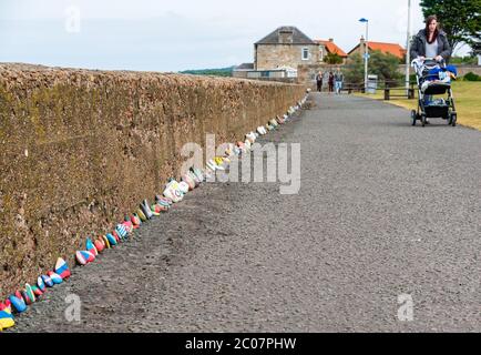 Port Seton, East Lothian, Scozia, Regno Unito. 11 Giugno 2020. Covid-19 simboli pandemici creati dalla gente del posto: Una linea lunga centinaia di piedi sul lungomare di pietre colorate e creative dipinte a mano con messaggi ispiranti. Sembra essere un fenomeno che si diffonde intorno alle città e villaggi della Lotia orientale. Una donna spinge un buggy sul sentiero oltre la linea di pietre Foto Stock