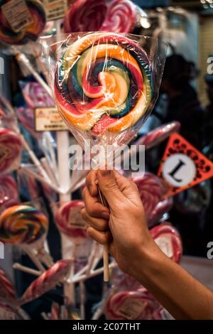 Mano femminile che tiene un lollipop di colore arcobaleno davanti ad un supporto con lollipop Foto Stock