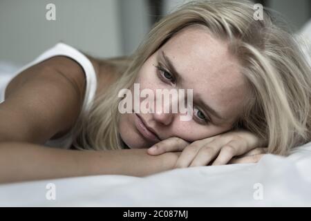 Triste bionda donna bianca che soffre di depressione e di interruzione del cuore che si trova sul suo letto a casa miserabile. Foto Stock
