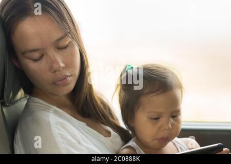 Un genitore e un bambino seduti su un addestramento che prende una pausa da una giornata impegnata di viaggio. Baby guardare la tv mentre la madre si riposa con gli occhi chiusi. Foto Stock