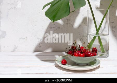 Ciliegie in una ciotola verde sotto foglie di monstera in un vaso di vetro su un rustico tavolo bianco e parete con ombre dure. Spazio di copia Foto Stock