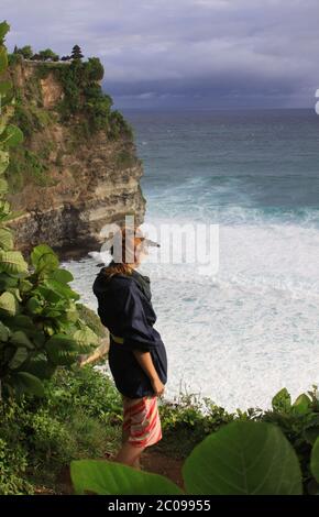 Donna a Uluwatu Bali in piedi e guardando le onde e l'oceano. La corrente del mare è forte e il tempio di Uluwatu si può vedere in lontananza. Foto Stock