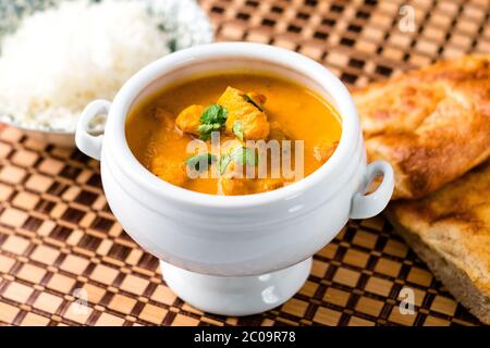 Delizioso piatto indiano di pollo al burro servito in una ciotola bianca con pane naan e riso sul lato. Il piatto viene posto su un tappetino di legno. Foto Stock