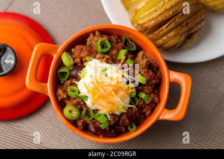 Ciotola di peperoncino con carne fresca fatta in casa con fagioli, panna acida, formaggio cheddar grattugiato e cipolle verdi. Servito con patate di asselback sul lato. Foto Stock