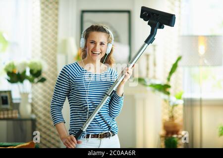 Ritratto di felice giovane donna in t-shirt a righe e pantaloni bianchi con aspirapolvere che ascolta la musica con le cuffie nella casa moderna sotto il sole Foto Stock