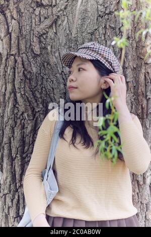 Attraente studente asiatico femminile del college guardando in lontananza mentre si appoggia contro UN albero Foto Stock