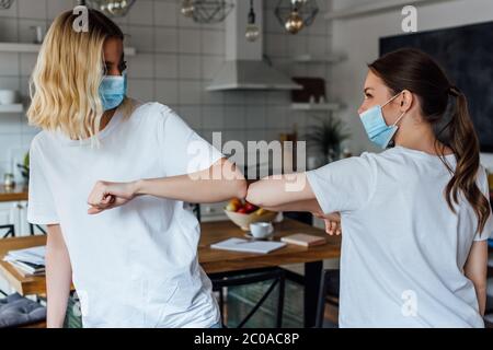 Sorelle in maschere mediche saluto con gomito in soggiorno Foto Stock