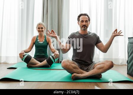 Uomo maturo meditando sul tappetino fitness vicino a sorridente moglie in soggiorno Foto Stock