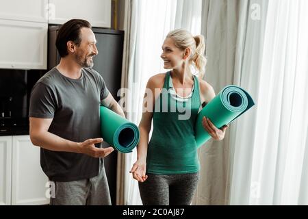 Uomo maturo sorridente che punta con la mano sul tappetino fitness vicino a bella moglie a casa Foto Stock