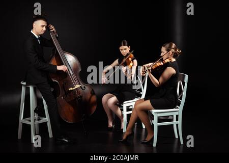 trio di musicisti professionisti che suonano musica classica su violini e contrabbasso su palcoscenico scuro Foto Stock