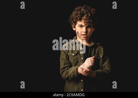 Bambino musulmano offeso con faccia sporca che tocca la mano ferita nella bendatura isolata su nero Foto Stock