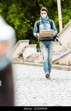 Fuoco selettivo del corriere in maschera medica che tiene le scatole della pizza mentre camminando sulla strada urbana Foto Stock