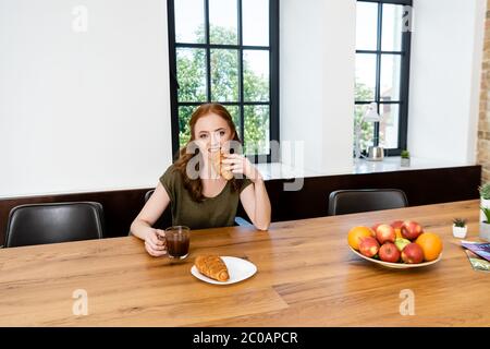 Donna dai capelli rossi che mangia croissant con una tazza di caffè a casa Foto Stock