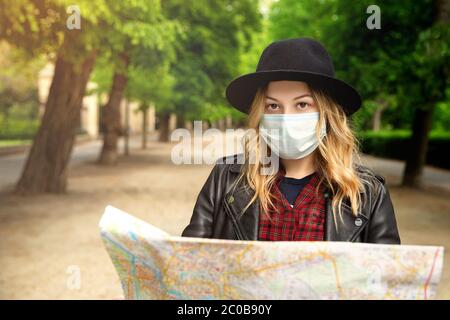 Una ragazza con una mappa della città è sulla strada. Guarda la macchina fotografica. Ha capelli biondi, in una giacca di pelle, in un cappello. Maschera facciale. Un nuovo concetto di viaggio in Foto Stock