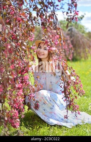 Una bella ragazza in un vestito blu e una paglia il cappello è seduto sotto un albero di mela rosa in fiore Foto Stock