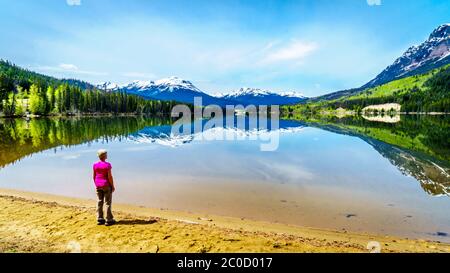 Donna anziana che guarda la riflessione della catena delle Montagne Rocciose sulla superficie liscia del lago Yellowhead nel Robson Provincial Park, Canada Foto Stock