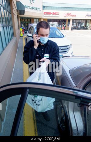 Uomo di consegna che indossa la maschera mettendo gli alimentari ordinati in auto nella corsia di prelievo che parla al telefono e mantenendo la distanza sociale. St Paul Minnesota, Minnesota, Stati Uniti Foto Stock