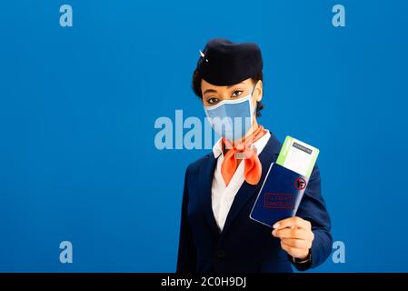 assistente di volo afroamericano in maschera medica con passaporto e biglietto aereo isolato su blu Foto Stock