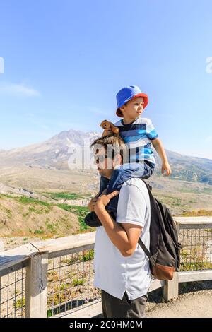Felice padre che porta sulle spalle il suo figlio piccolo che tiene un divertente giocattolo di chipmunk e guarda indietro con il Monte St. Helens in uno sfondo Foto Stock
