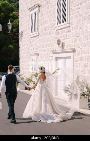 Una coppia di nozze cammina lungo la passeggiata sullo sfondo di una casa bianca vecchia, la sposa ondeggia un lungo velo, lo sposo tiene la sua mano. Bene Foto Stock