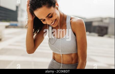 Donna runner con auricolari in piedi all'aperto e sorridente. Donna che si rilassa dopo un allenamento in città. Foto Stock