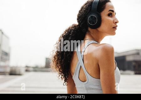 Donna fitness che indossa cuffie per ascoltare musica durante l'allenamento. Donna in abbigliamento sportivo che si prende pausa in piedi all'aperto e guardando via. Foto Stock