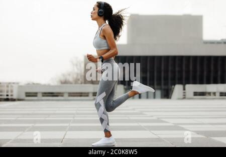 Foto a lunghezza intera di una donna che fa jogging in città. Donna che indossa abbigliamento sportivo e cuffie che corrono all'aperto. Foto Stock