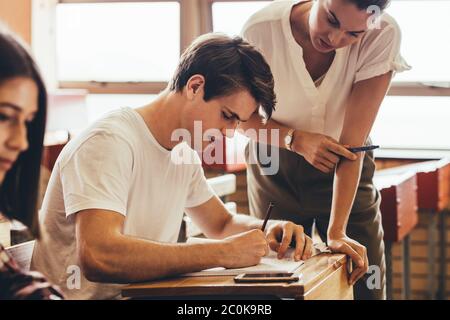 Studente universitario aiutato da una docente femminile durante la lezione. Giovane uomo che scrive nel suo libro con una insegnante femminile in piedi. Foto Stock