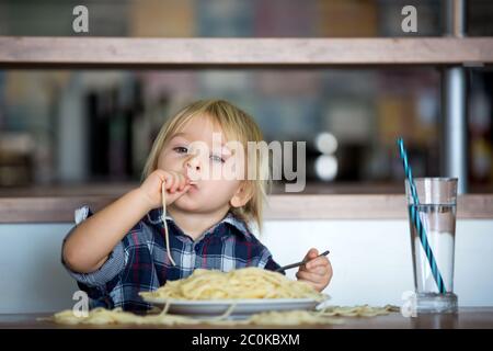 Bambino piccolo, bambino piccolo, mangiando gli spaghetti per pranzo e facendo l'alimentazione dell'amico dell'orso del teddy, pentola con gli spaghetti e i pomodori sul tavolo Foto Stock