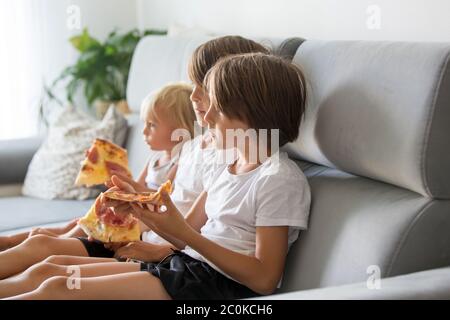 Bambini carini, seduti sul divano, mangiare pizza e guardare la TV. Bambino affamato che prende un morso dalla pizza durante una giornata di pizza party a casa Foto Stock