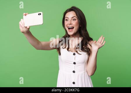 Ritratto di una bella ragazza bruna giovane eccitata con capelli lunghi che indossa abito estivo in piedi isolati su sfondo verde, prendendo un selfie Foto Stock