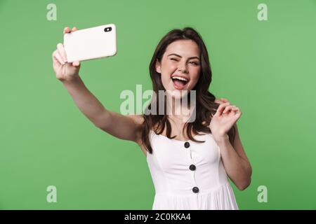 Ritratto di una bella ragazza bruna giovane eccitata con capelli lunghi che indossa abito estivo in piedi isolati su sfondo verde, prendendo un selfie, wink Foto Stock