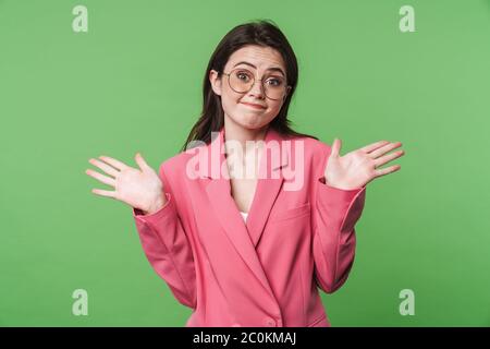 Ritratto di una bella giovane ed elegante donna confusa che indossa una giacca rosa in piedi isolata su sfondo verde, spalle ruggenti Foto Stock