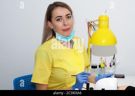 Cura delle unghie. Manicure in manicure. Il salone. Ragazza, manicurista, primo piano. Maschera e guanti di protezione. Mano del cliente con vernice sotto una lampada per Foto Stock