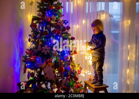 Bel ragazzo biondo, che decorano l'albero di natale con palle e corde di luce a casa Foto Stock