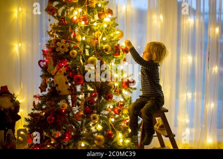 Bel ragazzo biondo, che decorano l'albero di natale con palle e corde di luce a casa Foto Stock