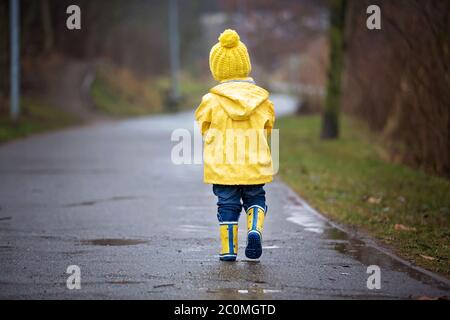 Bel ragazzo biondo divertente con anatre di gomma e ombrello colorato, saltando in puddddles e giocando sotto la pioggia, inverno Foto Stock