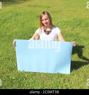 Una bella ragazza dai capelli lunghi sull'erba verde sul prato contiene un poster blu vuoto. Layout per la progettazione, posizionamento per il testo, spazio per la copia. Foto Stock