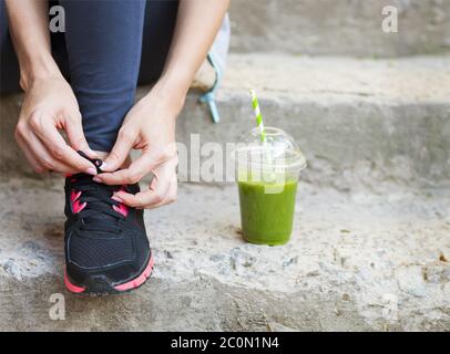 Coppa verde Detox frullato e donna allacciando scarpe da running prima dell'allenamento Foto Stock