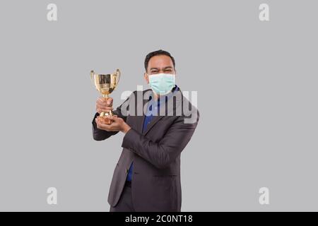 Businessman Holding Trophy indossare maschera medica. Uomo d'affari indiano in piedi con Trofeo in mani Foto Stock