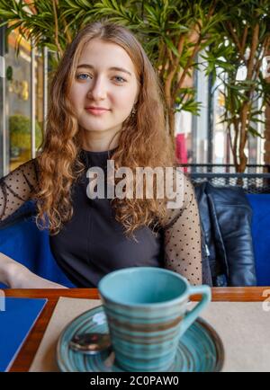 Una giovane donna molto bella, seduta dietro un gradino in un caffè e davanti a lei c'è una piccola ed elegante tazza con un motivo di cerchi di colori diversi. Foto Stock