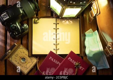 Notebook da viaggio, con passaporti, bussola, fotocamera fotografica, valigia e maschera chirurgica. Su tavolo di legno, illuminato da lampada a lume di candela. Foto Stock