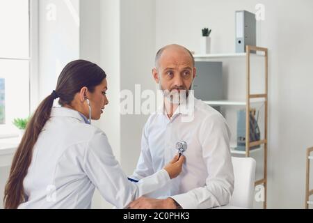 Donna medico cardiologo ascolta con stetoscopio l'uomo senatore paziente in clinica. Visita al medico in un ospedale medico. Foto Stock