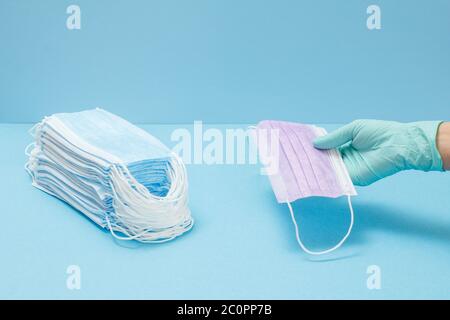 Una pila di maschere mediche protettive e una mano di un medico femminile in un guanto di nitrile che tiene una su sfondo blu. Dispositivi di protezione individuale ag Foto Stock